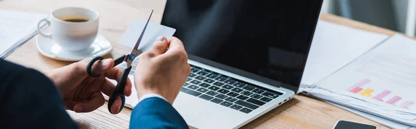 Plano panorámico de hombre de negocios con tijeras cerca de la tarjeta de crédito y el ordenador portátil - foto de stock