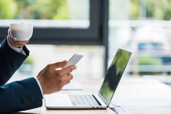 Vista ritagliata di uomo in possesso di una tazza di caffè e smartphone vicino al computer portatile — Foto stock