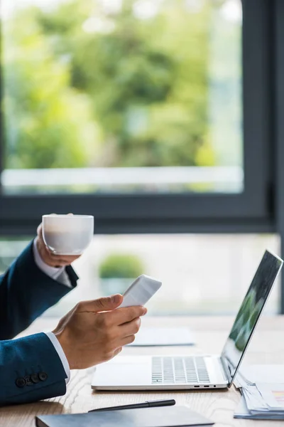 Vista recortada del hombre sosteniendo la taza y el teléfono inteligente cerca del ordenador portátil - foto de stock