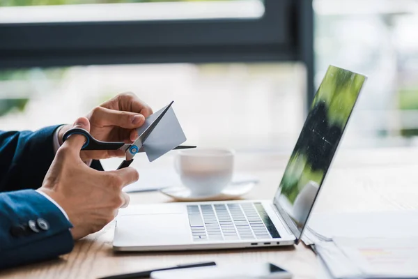 Vista recortada del hombre sosteniendo tijeras cerca de la tarjeta de crédito y gadgets en la mesa - foto de stock