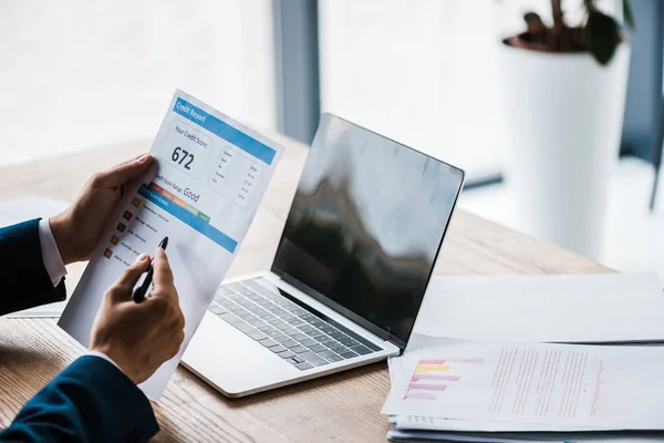 Vue recadrée de l'homme tenant du papier avec des lettres de rapport de crédit près d'un ordinateur portable avec écran blanc — Stock Photo