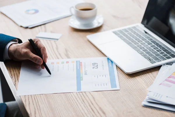 Cropped view of man holding pen near paper with credit report letters and cup — Stock Photo