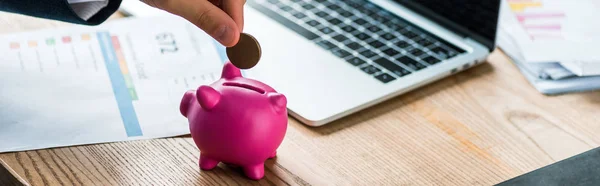 Tiro panorâmico de empresário colocando moeda no banco piggy rosa perto do laptop — Fotografia de Stock