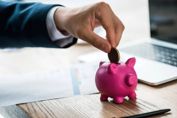 Cropped view of businessman putting coin into pink piggy bank near laptop — Stock Photo