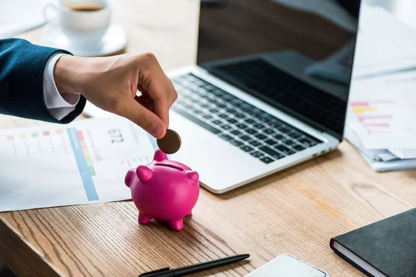 Vista recortada de hombre de negocios poner moneda en la alcancía rosa cerca de la computadora portátil y la taza con café - foto de stock