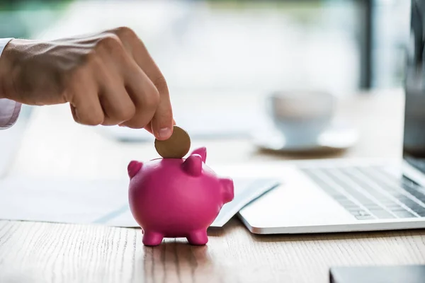 Vista recortada de hombre de negocios poner moneda en la alcancía rosa cerca de la computadora portátil - foto de stock