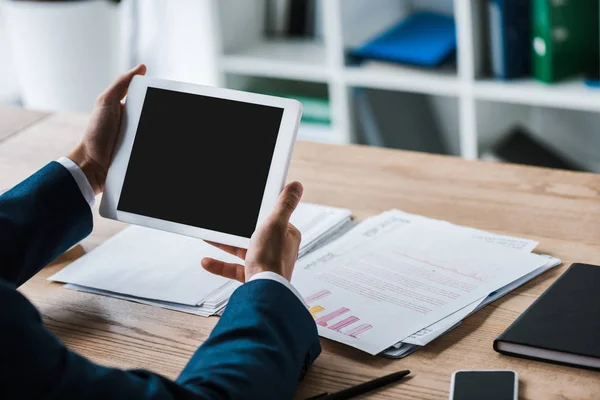 Vista recortada del empresario sosteniendo tableta digital con pantalla en blanco cerca de gráficos y gráficos en el escritorio - foto de stock