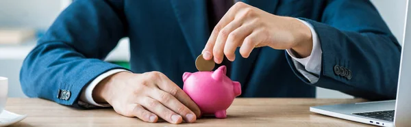 Panoramic shot of businessman putting metallic coin into piggy bank near laptop — Stock Photo