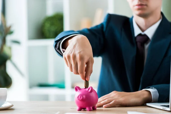 Cropped view of businessman putting metallic coin into piggy bank near laptop — Stock Photo