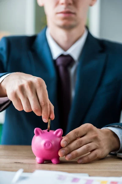 Selective focus of businessman putting metallic coin into piggy bank near laptop — Stock Photo