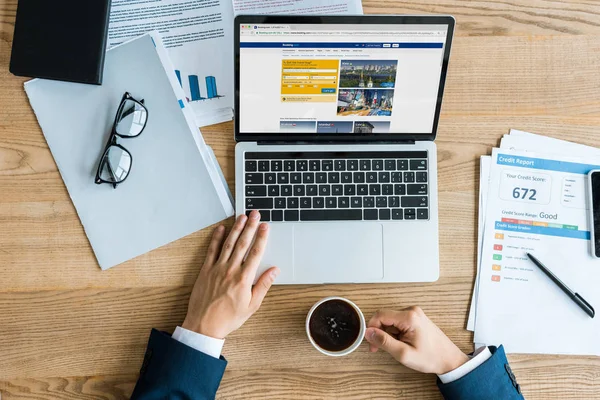 KYIV, UKRAINE - 8 JUILLET 2019 : vue du dessus de l'homme d'affaires tenant la tasse près de l'ordinateur portable avec le site Web de réservation à l'écran — Photo de stock