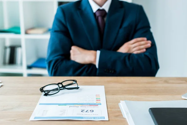 Enfoque selectivo de documento con informe de crédito cerca de gafas y hombre con brazos cruzados — Stock Photo