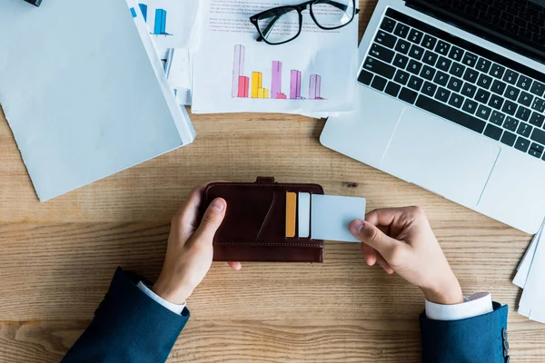 Vista superior del hombre sosteniendo la cartera con tarjetas de crédito cerca de la computadora portátil y gafas - foto de stock