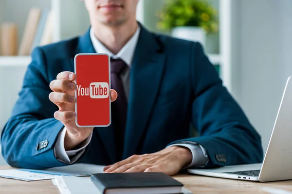 KYIV, UCRANIA - 8 de julio de 2019: enfoque selectivo del hombre que sostiene el teléfono inteligente con la aplicación de youtube en la pantalla cerca de la computadora portátil en la oficina - foto de stock