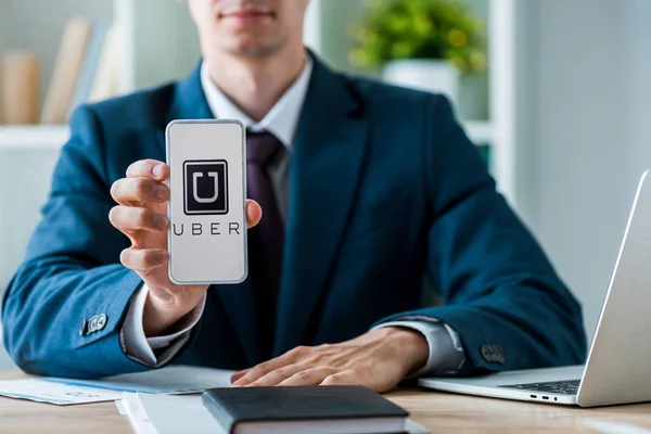 KYIV, UCRANIA - 8 de julio de 2019: enfoque selectivo del hombre que sostiene el teléfono inteligente con la aplicación uber en la pantalla cerca de la computadora portátil en la oficina - foto de stock