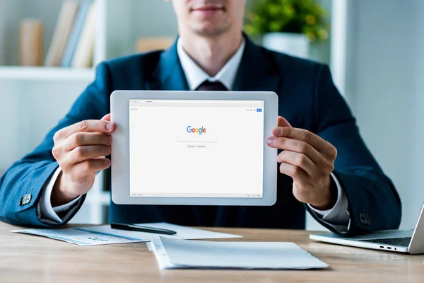 KYIV, UCRANIA - 8 de julio de 2019: enfoque selectivo del hombre que sostiene la tableta digital con la aplicación de Google en la pantalla cerca de la computadora portátil en la oficina - foto de stock