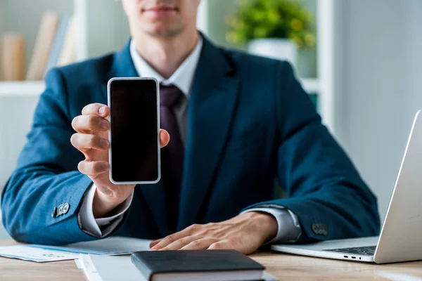 Enfoque selectivo del hombre que sostiene el teléfono inteligente con pantalla en blanco cerca de la computadora portátil en la oficina - foto de stock