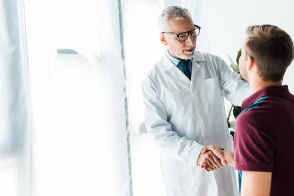 Happy doctor in glasses shaking hands with man in clinic — Stock Photo