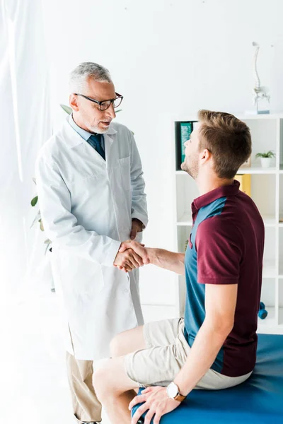 Heureux médecin dans des lunettes serrant la main avec jeune homme à la clinique — Photo de stock