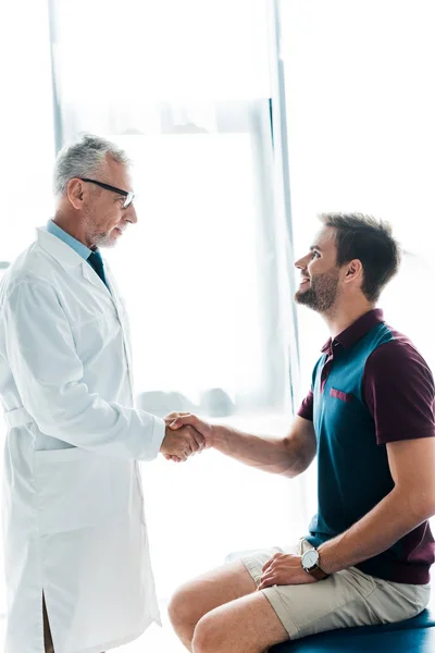 Happy doctor in glasses shaking hands with cheerful young man in clinic — Stock Photo