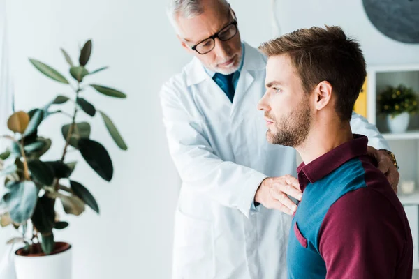 Selektiver Fokus des Arztes in Brille und weißem Mantel berührt gutaussehenden Mann in Klinik — Stockfoto