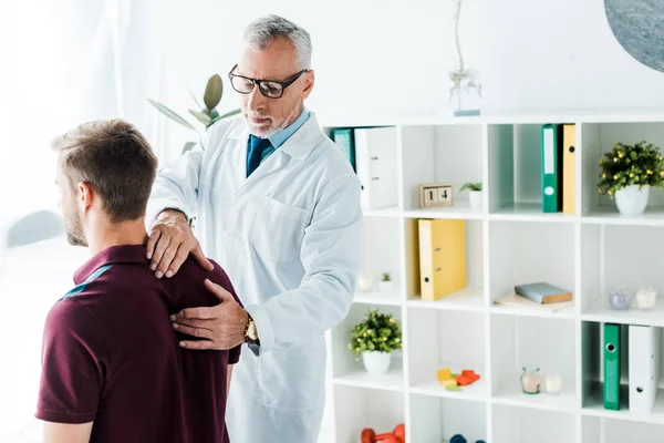 Хиропрактик в белом халате и очках, трогающих спину пациента в клинике — стоковое фото