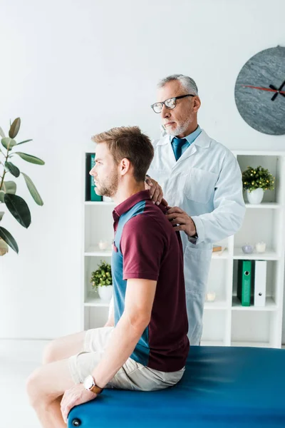 Chiropraktiker in weißem Mantel und Brille berührt Rücken eines gutaussehenden Patienten in Klinik — Stockfoto