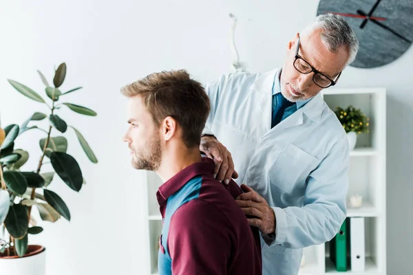 Quiropráctico barbudo en bata blanca y gafas tocando la espalda de un paciente guapo en la clínica - foto de stock