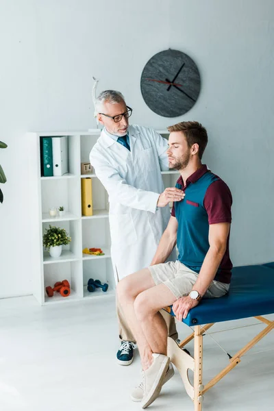 Bearded chiropractor in white coat and glasses touching back of patient with closed eyes — Stock Photo