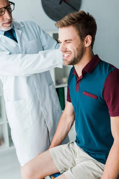 Хиропрактик в белом халате и очках трогает пациента, испытывающего боль в клинике — стоковое фото