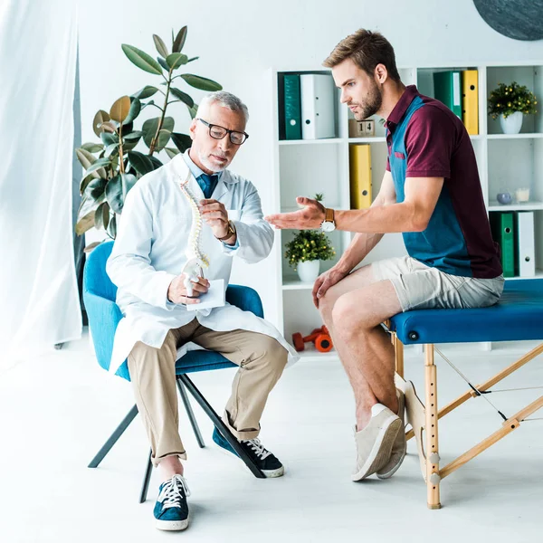 Beau barbu homme geste tout en regardant modèle colonne vertébrale près du médecin dans des lunettes — Photo de stock