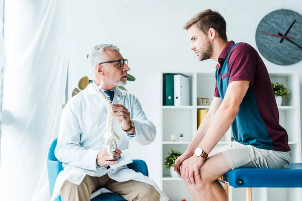 Bel homme barbu regardant médecin dans des lunettes tenant modèle de colonne vertébrale — Photo de stock