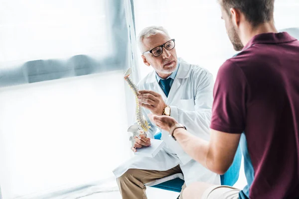 Selective focus of bearded doctor in glasses holding spine model near patient — Stock Photo