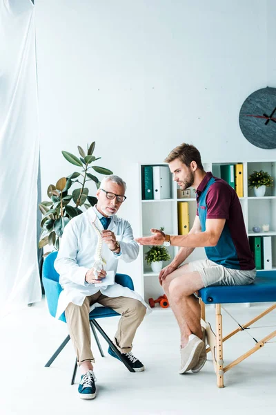 Bearded doctor in glasses holding spine model near patient gesturing in clinic — Stock Photo