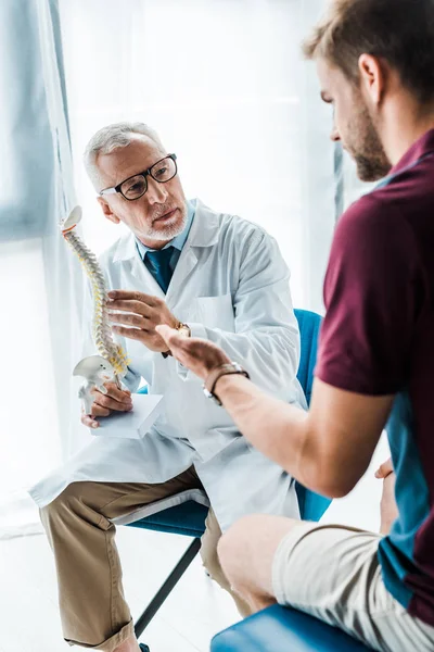 Foco seletivo do médico barbudo em óculos segurando modelo de coluna vertebral perto gestos paciente na clínica — Fotografia de Stock