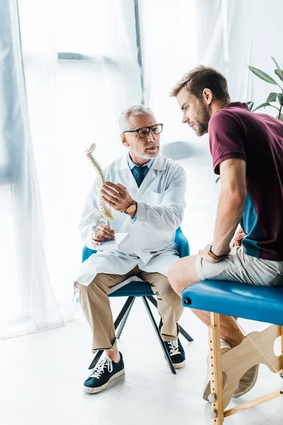Бородатый врач в очках с моделью позвоночника и глядя на пациента — стоковое фото