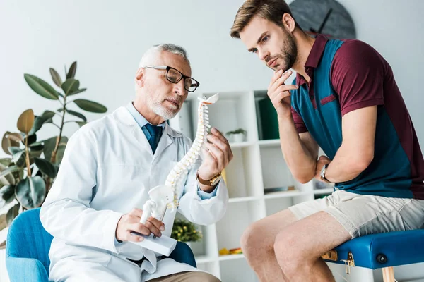 Nachdenklicher Mann schaut Wirbelsäulenmodel in weißem Mantel beim Arzt an — Stockfoto