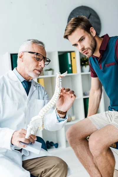 Médico barbudo sosteniendo modelo de columna vertebral cerca del paciente en el hospital - foto de stock