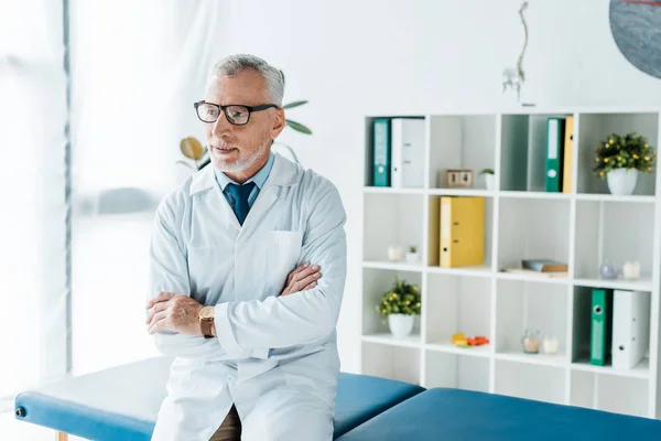 Doctor barbudo en gafas y abrigo blanco sentado en la mesa de masaje con los brazos cruzados - foto de stock