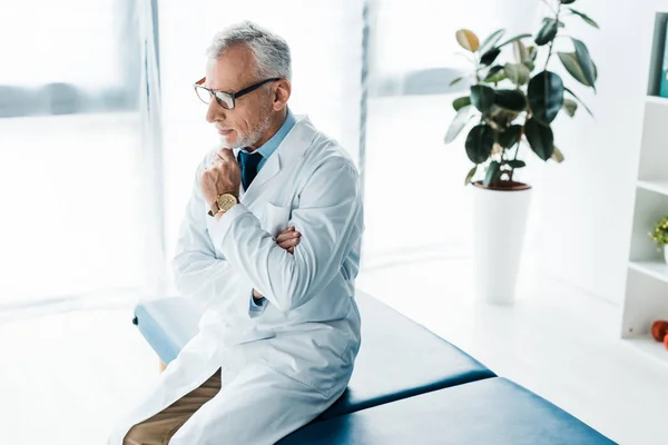 Médico barbudo pensativo en gafas y abrigo blanco sentado en la mesa de masaje y tocando la cara - foto de stock