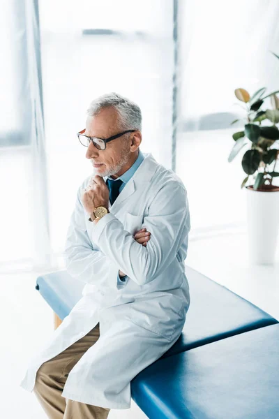 Médico barbudo reflexivo en gafas y abrigo blanco sentado en la mesa de masaje y la cara conmovedora - foto de stock