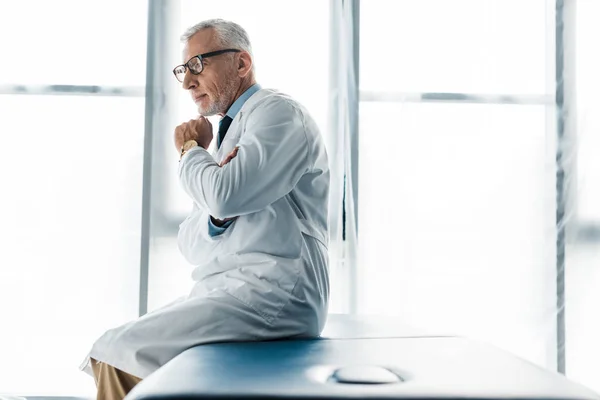 Thoughtful doctor in glasses and white coat sitting on massage table and touching face — Stock Photo