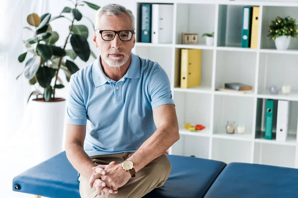 Bärtiger Mann mit Brille sitzt mit zusammengeballten Händen auf Massagetisch — Stockfoto