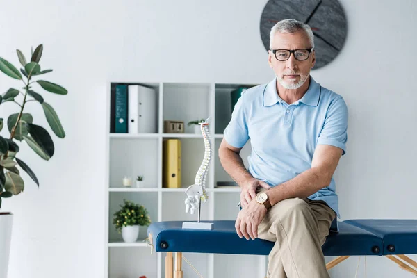 Barbudo hombre en gafas sentado en mesa de masaje cerca de la columna vertebral modelo - foto de stock