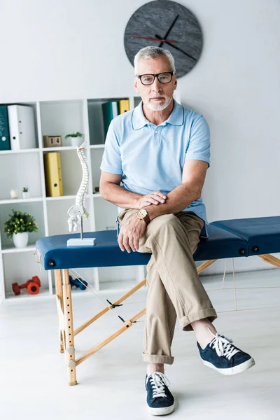Bärtiger Mann mit Brille sitzt auf Massagetisch in Klinik — Stockfoto
