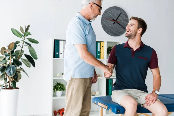 Hombre barbudo positivo en gafas estrechando la mano con paciente feliz en la clínica - foto de stock