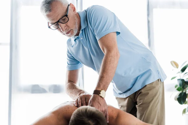 Бородатий хіропрактик в окулярах робить масаж без сорочки пацієнту на масажному столі — стокове фото