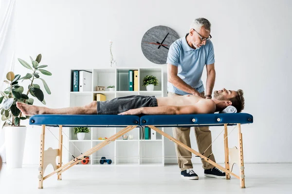 Бородатый хиропрактик делает массаж мужчине на массажном столе рядом с часами — стоковое фото