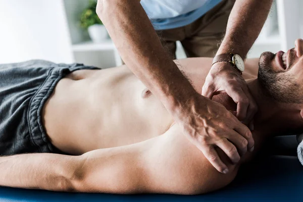 Cropped view of chiropractor doing massage to shirtless man with closed eyes suffering pain — Stock Photo