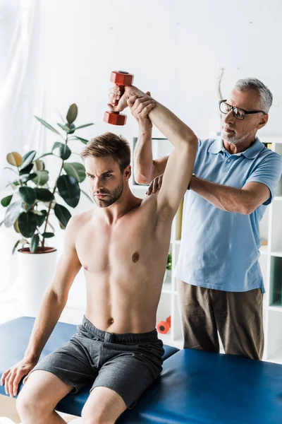Chiropraktiker mit Brille steht neben gutaussehendem Mann und übt mit Hantel in Klinik — Stockfoto